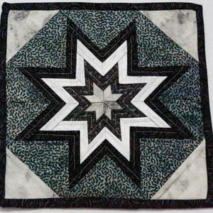 Puede incluir: Textil acolchado cuadrado con un diseño de estrella central. La estrella está compuesta por formas geométricas blancas, negras y grises, sobre un fondo de tela verde azulado estampada. Los bordes están acabados con un borde oscuro. Aproximadamente 25 cm x 25 cm.