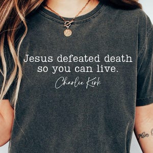 Puede incluir: Camiseta gris oscuro con el texto blanco: "Jesús venció la muerte para que puedas vivir. Charlie Kirk". La camiseta es de manga corta y cuello redondo. La imagen también incluye texto PNG y SVG.