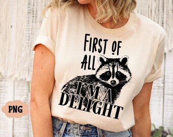 May include: A beige t-shirt with a black graphic of a raccoon and the text "First of all I'm a Delight".