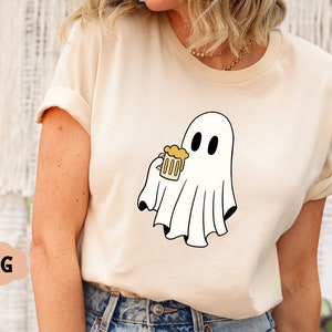 Pu&ograve; includere: Una maglietta bianca con un fantasma dei cartoni animati che tiene una boccale di birra gialla. Il fantasma ha due occhi neri e un corpo bianco a forma di lenzuolo.