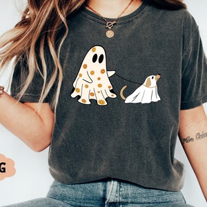 Puede incluir: Una camiseta gris con un gr&aacute;fico de un fantasma que lleva un estampado floral y sostiene una correa atada a un perro fantasma. El texto "More Bean" est&aacute; escrito en la manga derecha de la camiseta.