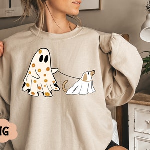 Puede incluir: Una sudadera beige con un estampado gr&aacute;fico de un fantasma con un patr&oacute;n floral y un perro con un disfraz de fantasma. El fantasma sostiene una correa atada al perro.