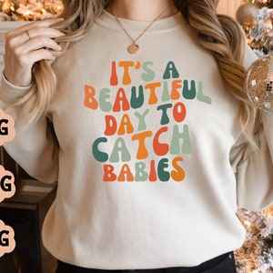 Puede incluir: Una sudadera de color crema con el texto "It's a beautiful day to catch babies" en letras coloridas de estilo retro.