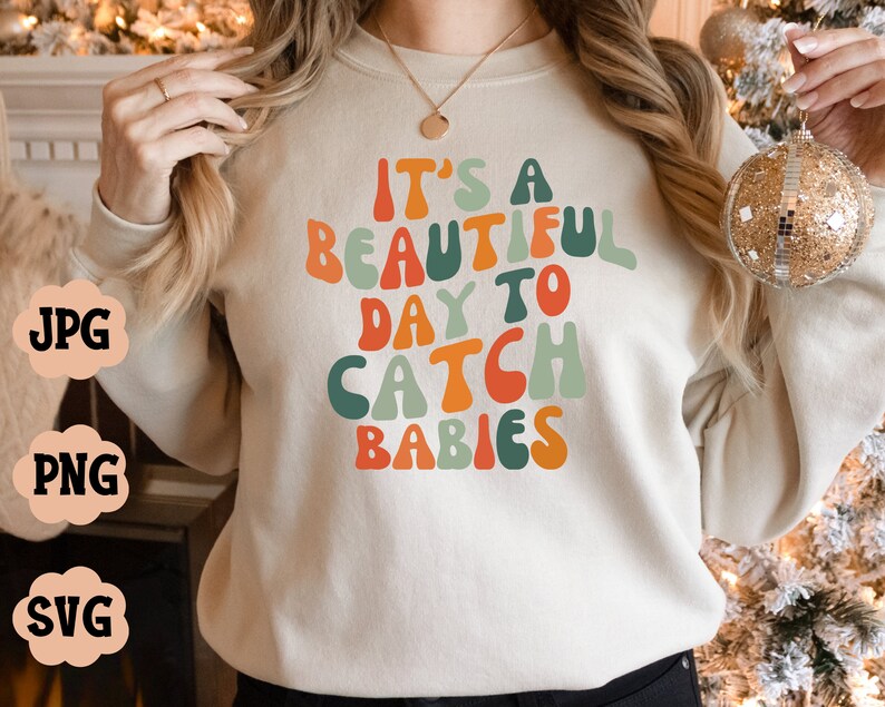Puede incluir: Una sudadera de color crema con el texto "It's a beautiful day to catch babies" en letras coloridas de estilo retro.