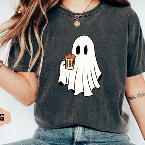 Pu&ograve; includere: Una maglietta grigia con un fantasma bianco che tiene un bicchiere di liquido arancione. Il fantasma ha due occhi neri e una piccola bocca. Il testo "More Beer" &egrave; scritto sulla manica destra della maglietta.