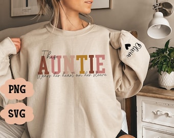 May include: A beige sweatshirt with the text "This Auntie wears her heart on her sleeve" in pink, brown, and gold lettering. The sleeve has a black heart with the word "Auntie" written inside.