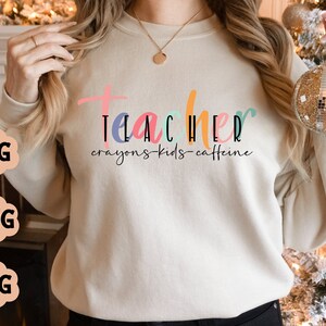 May include: A white sweatshirt with the text "Teacher crayons-kids-caffeine" in colourful letters.