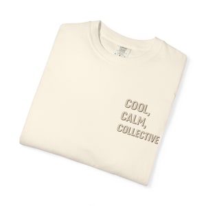 May include: A cream-colored crewneck t-shirt featuring the text COOL, CALM, COLLECTIVE embroidered in a light brown, block-style font on the lower right side. The shirt has a visible interior neck label with size L and brand information.