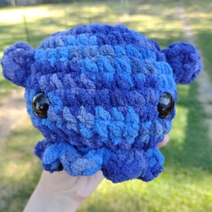 May include: A blue and gray crocheted octopus plush toy with black eyes. The octopus is being held in a hand.