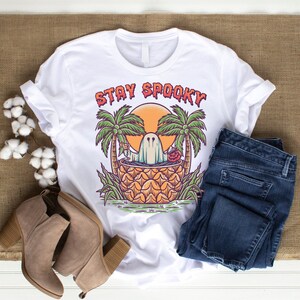 May include: White t-shirt with a graphic of a ghost in a pineapple with palm trees and the text "Stay Spooky".