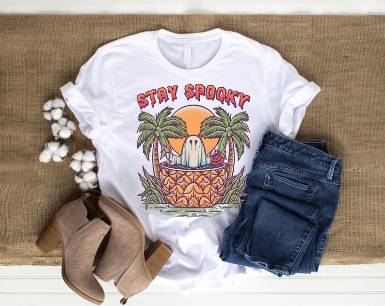 May include: White t-shirt with a graphic of a ghost in a pineapple with palm trees and the text "Stay Spooky".