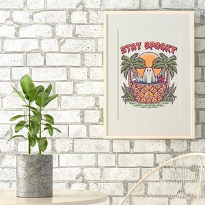 May include: A framed print with a white background and a colorful illustration of a ghost in a pineapple with palm trees and the text "Stay Spooky".