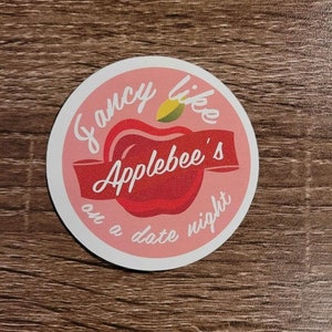 Puede incluir: Una pegatina redonda de color rosa y rojo con el texto "Fancy like Applebee's on a date night" y una manzana roja con una hoja verde.