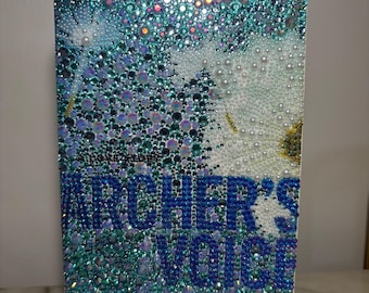 May include: A blue and white book cover with the title "Archer's Voice" and the author's name "Mia Sheridan" in rhinestones. The cover features a design of white dandelions with iridescent blue and green accents.