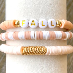 Peut inclure: Trois bracelets avec des perles blanches et pêche. Le bracelet du haut a des perles blanches avec le mot "FALL" écrit en lettres dorées. Le bracelet du milieu a des perles blanches et pêche alternées. Le bracelet du bas a des perles pêche avec un accent en métal doré.