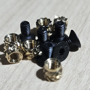 May include: A collection of black screws and gold-colored threaded inserts. The screws have a hex head design, while the inserts have a fluted exterior. The hardware is likely for furniture or woodworking projects.