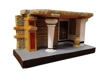 Pode incluir: Um modelo arquitetónico em miniatura de uma estrutura com paredes de textura de pedra, um telhado plano e uma coluna central branca. A fachada apresenta detalhes em amarelo e vermelho, uma pequena urna bege sobre base cinzenta e um painel.