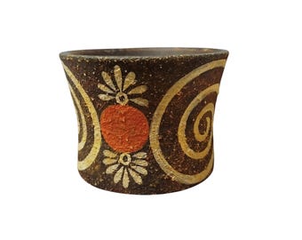 Pode incluir: Jarro cilíndrico de cerâmica artesanal com base castanha escura. O jarro apresenta um padrão repetido de espirais creme, círculos laranja e desenhos florais brancos. O jarro é provavelmente usado para fins decorativos.