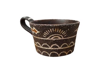 Pode incluir: Caneca de cerâmica marrom escura feita à mão com uma alça curva. A caneca apresenta desenhos geométricos pintados em branco e dourado, incluindo um padrão de raios de sol e padrões de vieiras ao redor da borda. O interior é marrom claro.