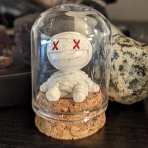 May include: A small, white mummy figurine with red 'X' eyes, encased in a glass dome. The mummy sits on a cork base. The background includes dark objects and a stone.