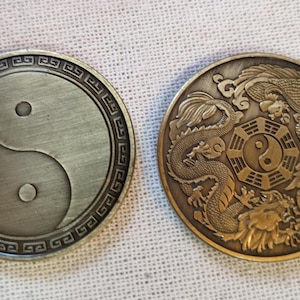 May include: Two round, metal coins with a yin yang symbol on one side and a dragon and phoenix design on the other. The yin yang symbol is in black and white, while the dragon and phoenix are in gold.
