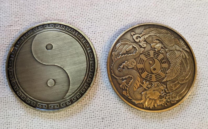 May include: Two round, metal coins with a yin yang symbol on one side and a dragon and phoenix design on the other. The yin yang symbol is in black and white, while the dragon and phoenix are in gold.
