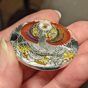 以下が含まれることがあります： カラフルな鳳凰のデザインが施された銀色の円形ペンダント。鳳凰は赤、黄、青、緑のアクセントが施されています。ペンダントは複雑なパターンとテクスチャのある表面で装飾されています。
