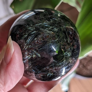 May include: A polished spherical gemstone featuring a clear, transparent outer layer with dark, deep brown and black inclusions. Inside the sphere, thin, needle-like metallic green and silver mineral fibers are visible, creating a reflective pattern.