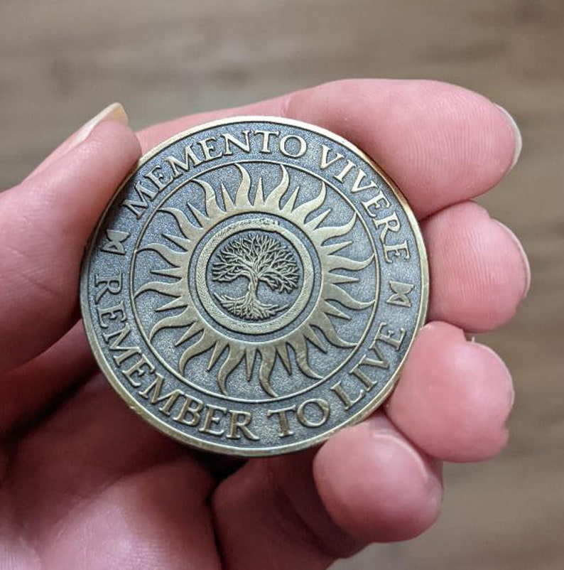 Peut inclure: Une pi&egrave;ce ronde de couleur bronze avec les mots "Memento Vivere Remember To Live" inscrits sur le bord. Le centre de la pi&egrave;ce pr&eacute;sente un soleil stylis&eacute; avec un arbre &agrave; l'int&eacute;rieur.