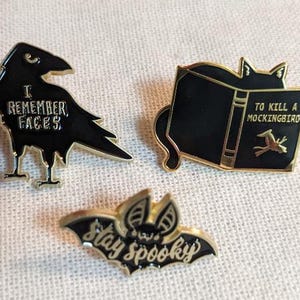 May include: Three black enamel pins with gold accents. The first pin features a black crow with the text "I remember faces". The second pin features a black cat reading a book with the text "To Kill a Mockingbird". The third pin features a black bat with the text "Stay Spooky".