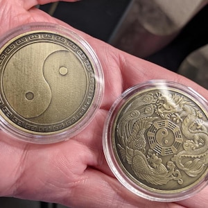 May include: Two round, gold-colored metal coins with decorative designs. The first coin features a yin yang symbol with a black and white design. The second coin features a dragon and a phoenix with a yin yang symbol in the center.