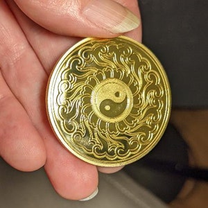 以下が含まれることがあります： 中心に陰陽のシンボルが描かれた、複雑なデザインの金色の金属コイン。コインは縁が高く、表面は滑らかです。