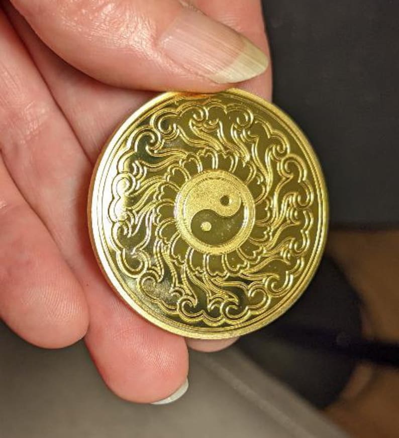 以下が含まれることがあります： 中心に陰陽のシンボルが描かれた、複雑なデザインの金色の金属コイン。コインは縁が高く、表面は滑らかです。