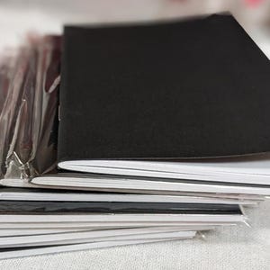 May include: A stack of small notebooks with black covers and white pages. Some notebooks are individually wrapped in clear plastic. The notebooks are stacked on top of each other, with the top notebook open.
