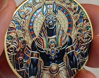 May include: A round, gold-toned coin featuring an Anubis design. The Anubis figure is depicted with a black body and a jackal head, surrounded by a colorful stained-glass style background. The coin has intricate details and a polished finish.