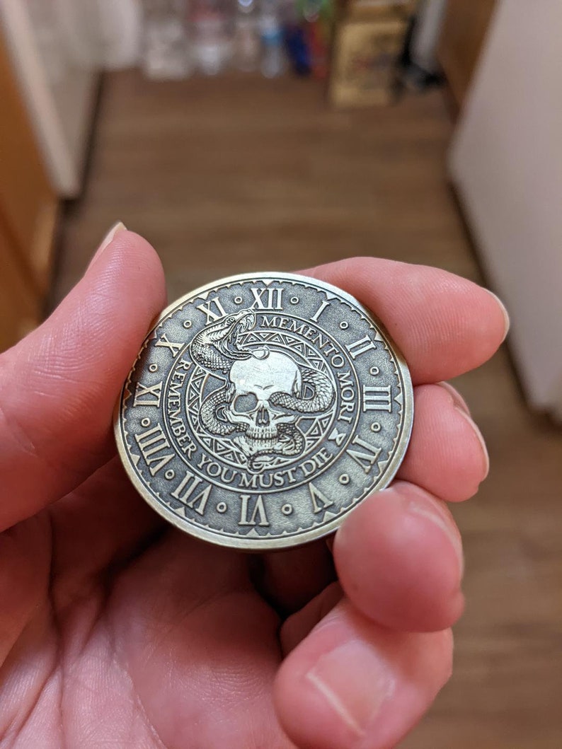 Peut inclure: Une pi&egrave;ce ronde de couleur laiton avec un motif de cr&acirc;ne et de serpent. La pi&egrave;ce comporte des chiffres romains autour du bord et le texte "Memento Mori Remember You Must Die".