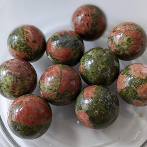 May include: A collection of ten polished unakite spheres. The spheres display a mix of colors, including green, red, and pink, with a speckled pattern. The spheres are arranged in a clear glass bowl.