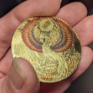 以下が含まれることがあります： 炎から昇るフェニックスの細密な画像が描かれた金色のコイン。フェニックスは赤、オレンジ、紫の羽を持ち、渦巻く模様に囲まれています。