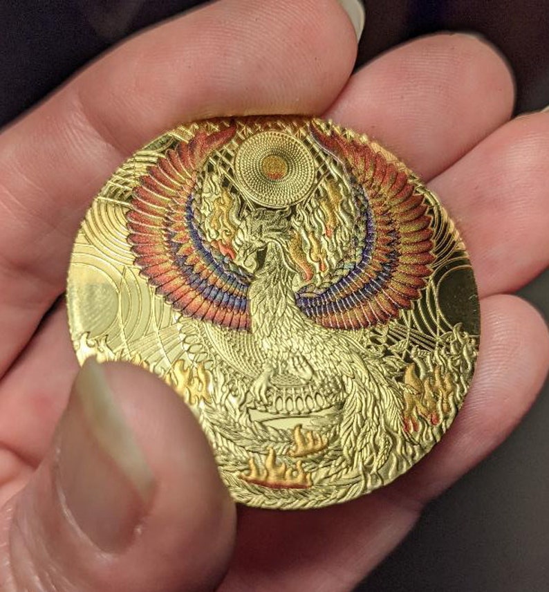 以下が含まれることがあります： 炎から昇るフェニックスの細密な画像が描かれた金色のコイン。フェニックスは赤、オレンジ、紫の羽を持ち、渦巻く模様に囲まれています。