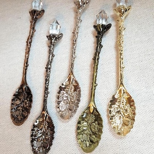 May include: Five decorative metal spoons with textured branch-like handles and clear crystal points at the tips. Each spoon bowl features an embossed floral and leaf pattern. The set includes finishes in copper, dark gunmetal, silver, brass, and gold.