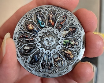 May include: A silver-colored zodiac wheel with a sun in the center and twelve zodiac signs around the outside. The signs are illustrated with colorful images of the zodiac animals. The signs are: Aquarius, Pisces, Aries, Taurus, Gemini, Cancer, Leo, Virgo, Libra, Scorpio, Sagittarius, and Capricorn.
