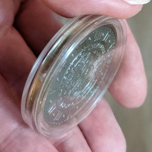Peut inclure: Une pi&egrave;ce d'argent avec des chiffres romains et un motif au centre, enferm&eacute;e dans un &eacute;tui de protection en plastique transparent.