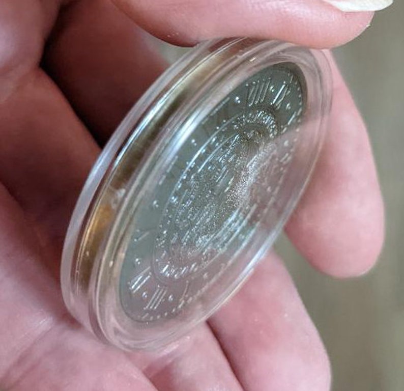 Peut inclure: Une pi&egrave;ce d'argent avec des chiffres romains et un motif au centre, enferm&eacute;e dans un &eacute;tui de protection en plastique transparent.