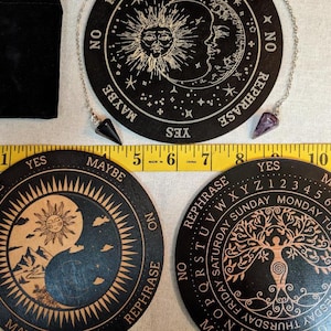 May include: A collection of divination tools, including three black wooden planchette boards with celestial and tree of life designs, two crystal pendulums, and a black velvet bag. A yellow measuring tape is also present.