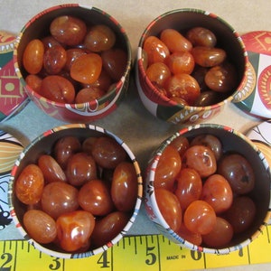 May include: Four small, round tins filled with polished, orange-brown carnelian stones. The tins have decorative rims with red, green, and yellow patterns. The stones are smooth and vary in size and shade.