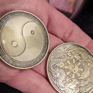May include: Two gold-colored metal coins with a yin yang symbol on one side and a dragon and phoenix design on the other. The yin yang symbol is black and white, and the dragon and phoenix are in a stylized design.