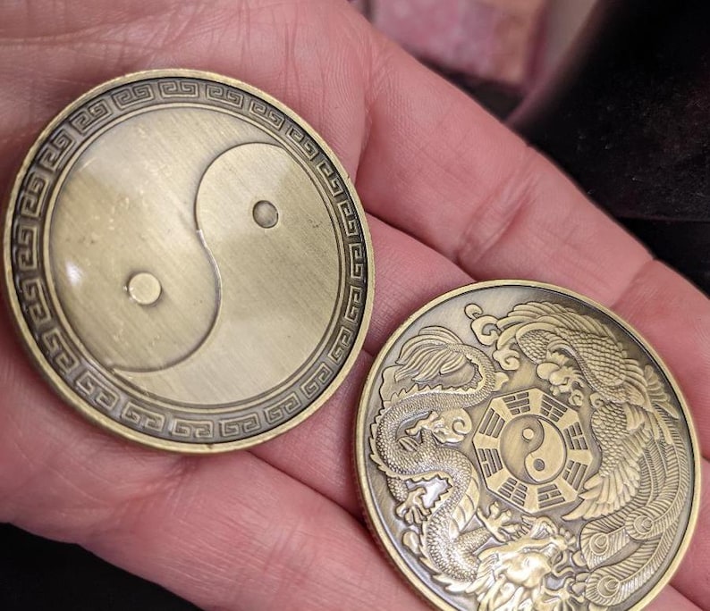May include: Two gold-colored metal coins with a yin yang symbol on one side and a dragon and phoenix design on the other. The yin yang symbol is black and white, and the dragon and phoenix are in a stylized design.