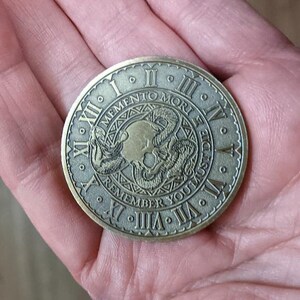 Peut inclure: Une pi&egrave;ce ronde de couleur laiton avec un motif de cr&acirc;ne et de serpent. La pi&egrave;ce comporte des chiffres romains autour du bord et le texte "Memento Mori Remember You Must Die".