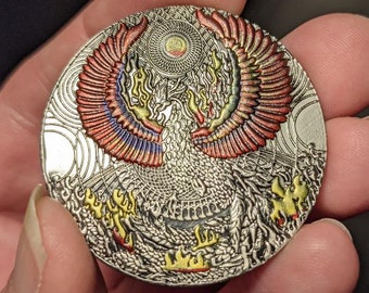 May include: A silver circular coin with a detailed design of a phoenix rising from flames. The phoenix has red, orange, yellow, and blue wings. The background is silver with a circular pattern.