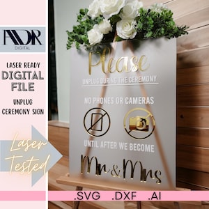 Pode incluir: Uma placa branca fosca com letras douradas que diz "Por favor, desligue durante a cerimônia. Sem telefones ou câmeras até que nos tornemos Sr. e Sra.". A placa está em um cavalete de madeira e tem um arranjo floral no topo.